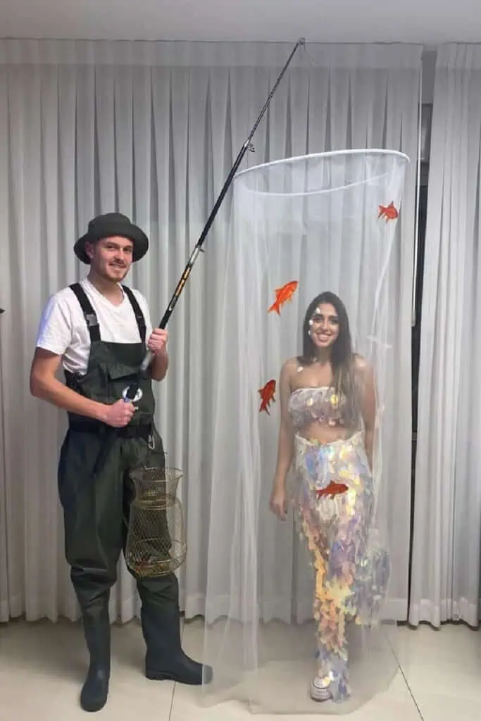Funny couples Halloween costume: a fisherman in green waders and boots holding a fishing rod and basket stands next to his &ldquo;biggest catch,&rdquo; a woman inside a tall tulle net wearing iridescent sequin &ldquo;fish scale&rdquo; outfit with orange goldfish decorations.
