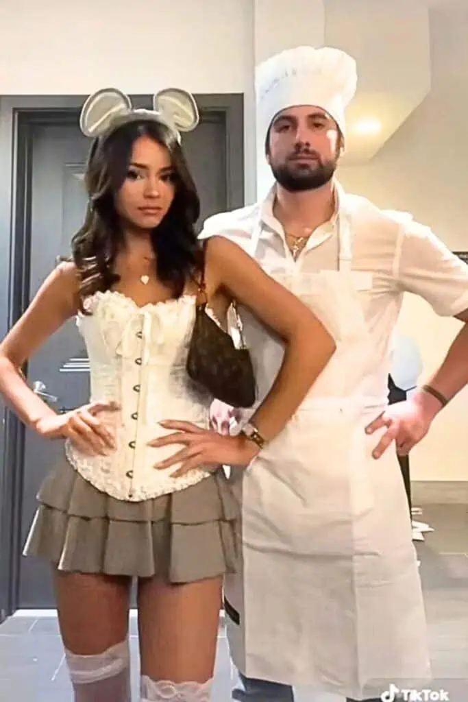 Couple dressed as Remy and Linguini from Ratatouille for Halloween, with the woman wearing a white corset and mouse ears and the man in a chef&rsquo;s outfit and apron, posing indoors in a trendy DIY couples costume cute.